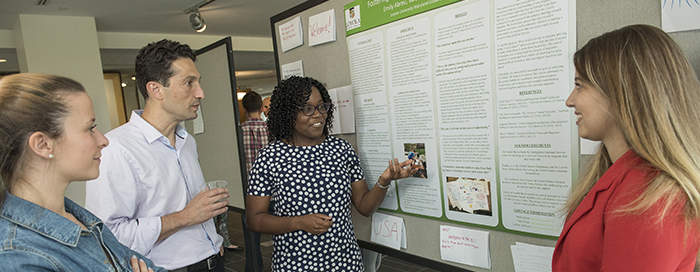 A female student pointing to her research presentation poster as she speaks with three other individuals
