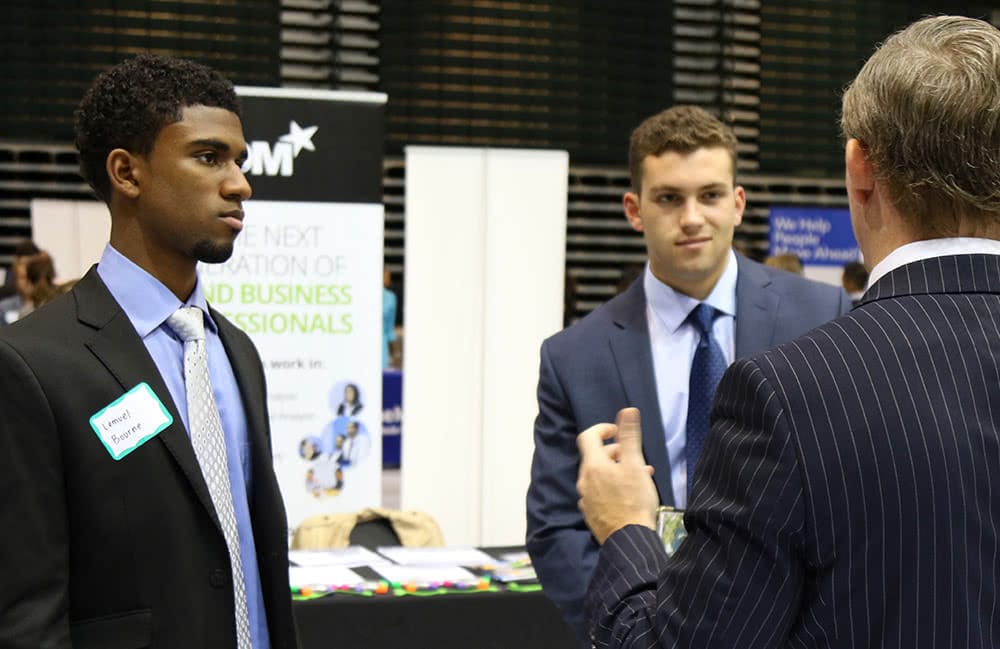 Two students wearing business suits talk to a recruiter at the career fair