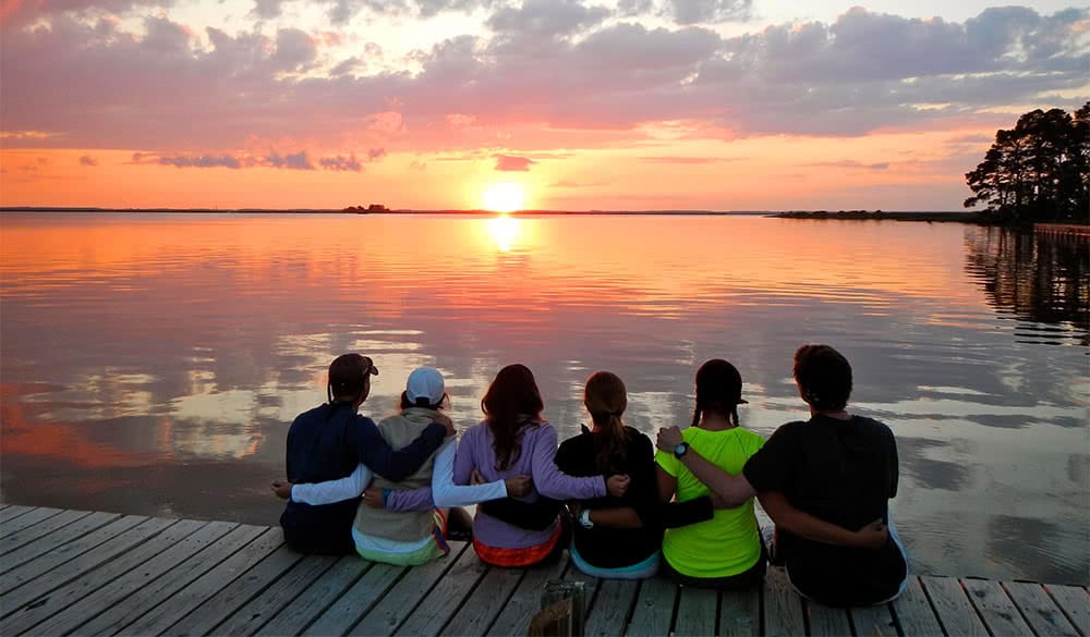 Students sitting on the edge of a pier with their arms around each other and looking out at a colorful sunset on the water