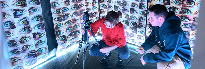 Students crouching with a camera in a brightly lit room filled with photographs of closeup angles of eyes