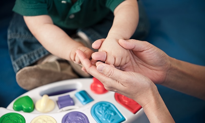 Adult hands holding a toddler's hands with a learning toy underneath on the floor