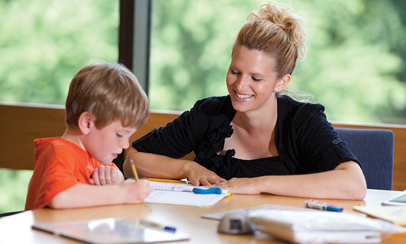 A young child writes on paper, sitting next to a smiling woman