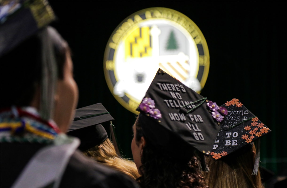 Students wearing decorated graduation caps looking at a stage with the Loyola seal in the background