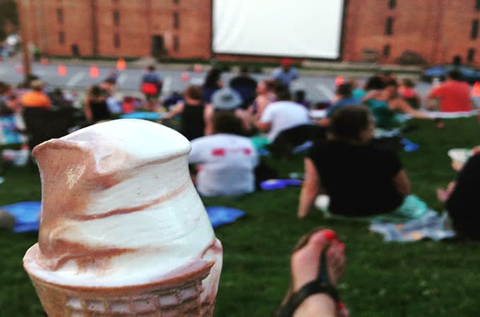 First-person view of a person holding ice cream while sitting in a grassy field watching an outdoor movie