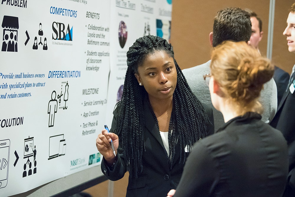 A student standing in front of a presentation board speaking to visitors at a Pitch Competition