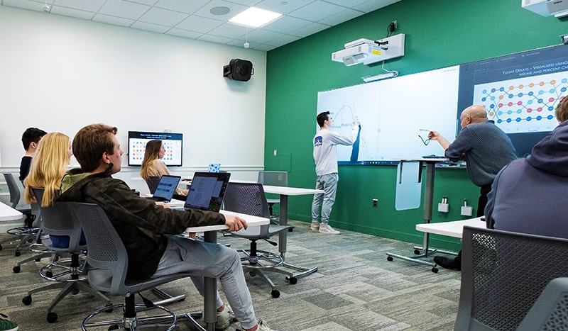 A student stands at the front of the Data Visualization Lab classroom, drawing a data visualization while classmates and his professor watch