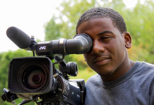 A Black student holds a camera up to his face as he films.