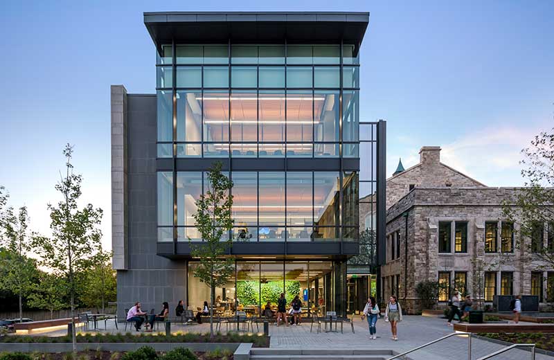 Loyola community members sitting and walking around the ambiently lit glass facade of the Fernandez Center at dusk