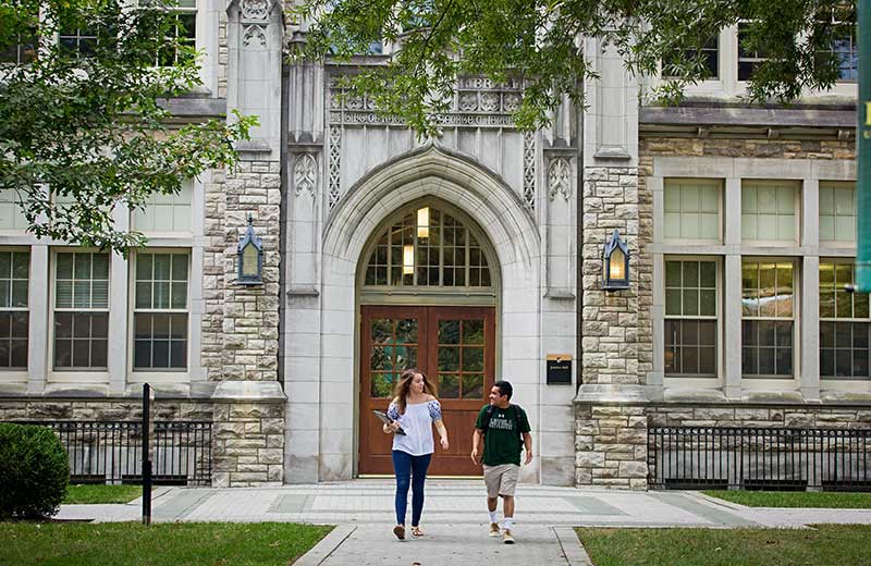 Two students chatting and walking away from the large wooden front doors of an ornate stone campus building