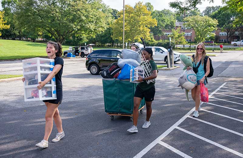 Three student laughing while carrying arms full of bedding and move-in supplies