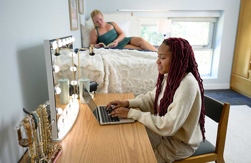 Two roommates together in their room, one reading on her bed and one using a laptop at her desk