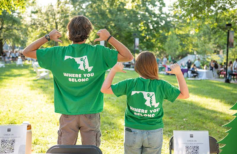 Two students with their backs to the camera, pointing to their shirts that say: Where You Belong