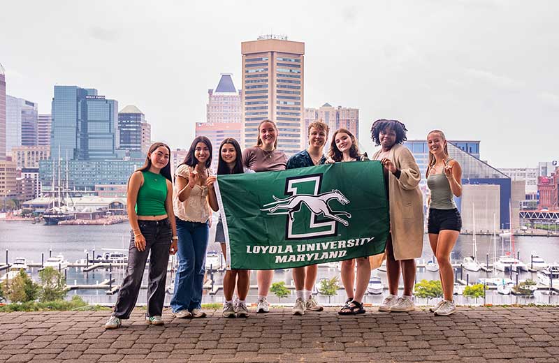 Group of students holding up a Loyola flag and making greyhound hand signs on a hill that overlooks Baltimore City