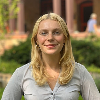 Camille, smiling by the green front garden of the Humanities building