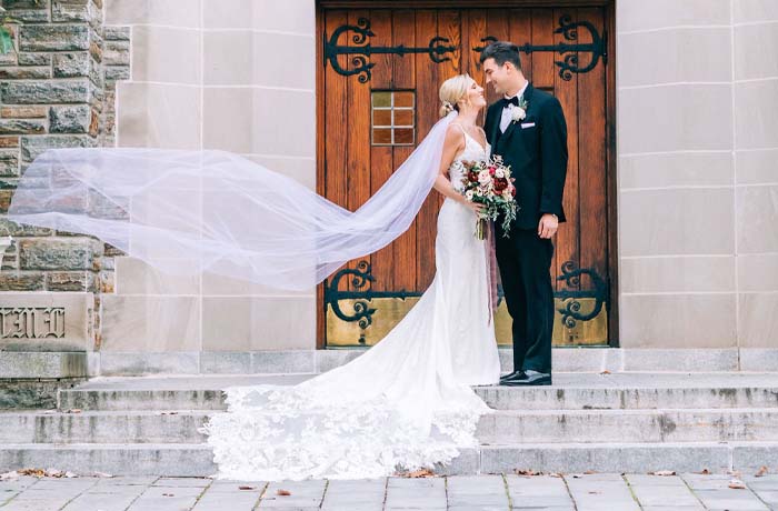 A recently married couple pose for a photo in front of the doors to Loyola's Alumni Memorial Chapel while the wind lifts the brides veil elegantly in the wind