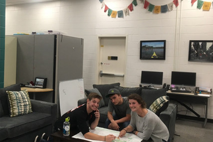Three students smiling and posing in their OAE office while using a map to plan out a trip