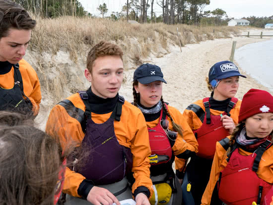 Group of students in waterproof jumpsuits and life jackets, looking out onto the water as they discuss their kayak trip