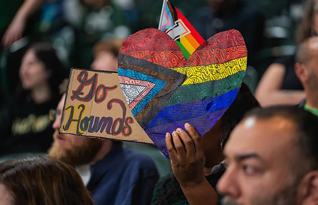 A student in a crowd at an athletics game holding a rainbow-colored heart that says: Go Hounds