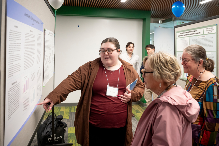 Graduate student gestures to poster for two visitors while presenting at Emerging Scholars Loyola University Maryland