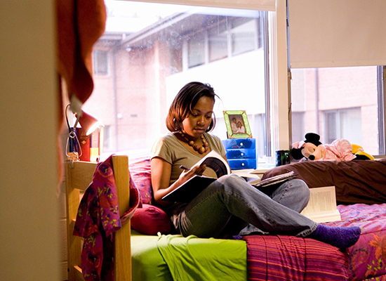 A student sits on a bed in their dorm room reading a book