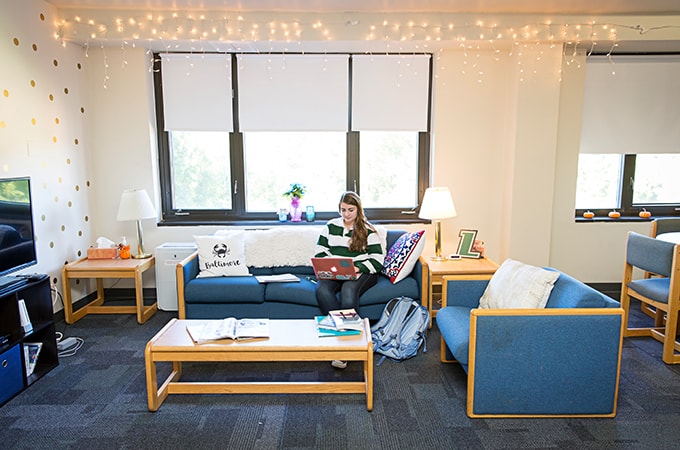 A student seated on a couch in their dorm room works on a laptop