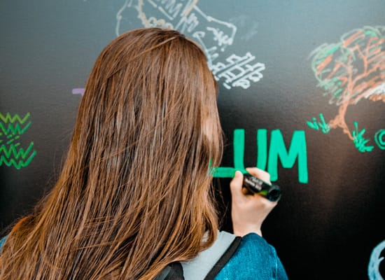 A student writing 'LUM' on a chalkboard, while other doodles can be seen on the chalkboard