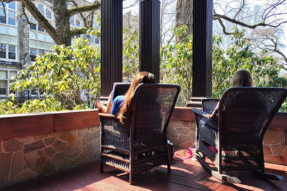 Two students sit in rocking chairs on the porch of the Humanities Building while working on their laptops