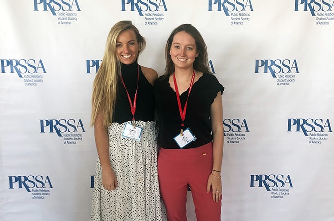 Isabelle Garrity and Julia Mulry posing for a photograph in front of a PRSSA step and repeat banner