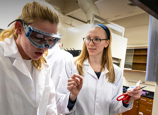Students wearing white lab coats and safety goggles in a lab