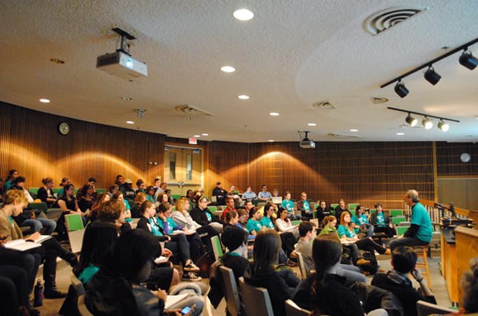 A professor sitting on a stool in front of a crowded classroom