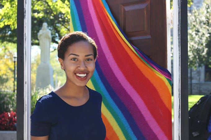 A female student standing outside in a doorframe with a rainbow flag draped on the door