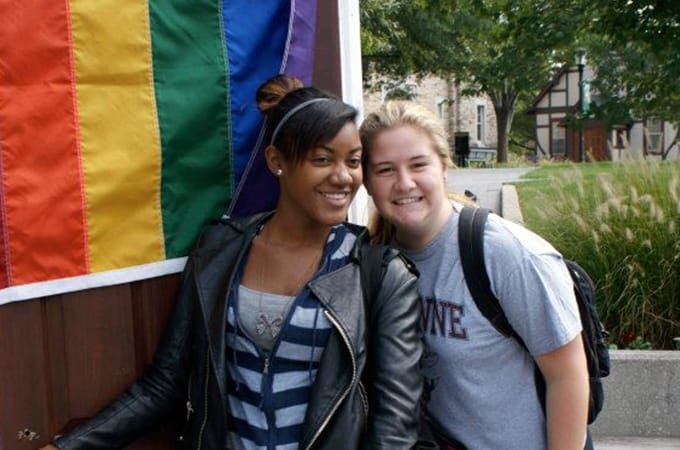 A male student and a faculty advisor standing outside in a doorframe with a rainbow flag draped on the door