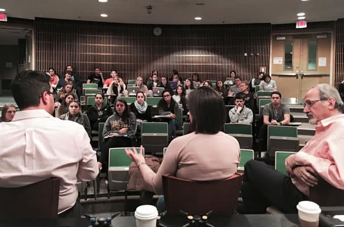 Three panelists seen from behind talking in front of a classroom full of students