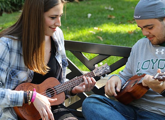 Two students playing ukuleles while sitting on a bench outside 