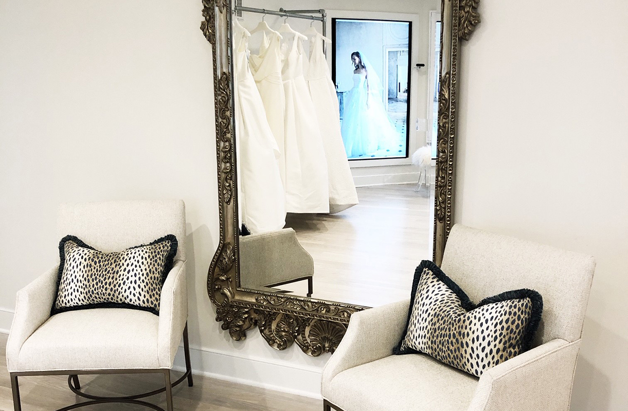 Two chairs flank a large, ornate mirror. In the distant double reflection, a client tries on a dress.