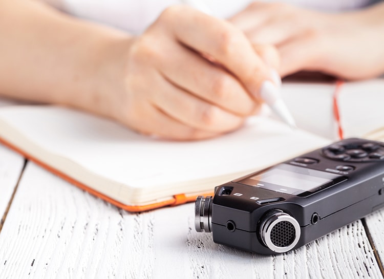 Photo of a person writing in a notebook with a voice recorder in the foreground