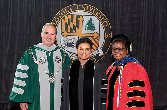 Terry Sawyer, Anisya Fritz, and Cheryl Moore-Thomas dressed in regalia posing in front of the Loyola seal