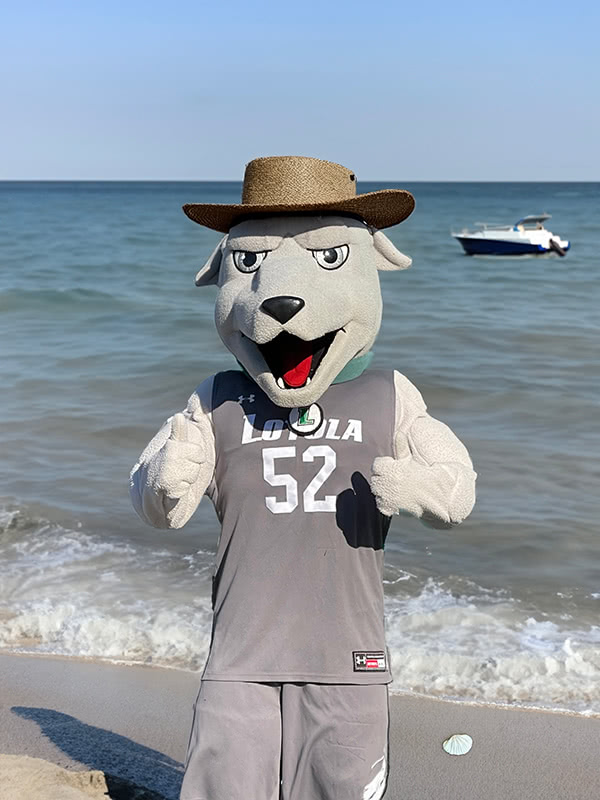 Greyhound Iggy mascot standing in front of an AI-generated beach wearing an AI-generated hat