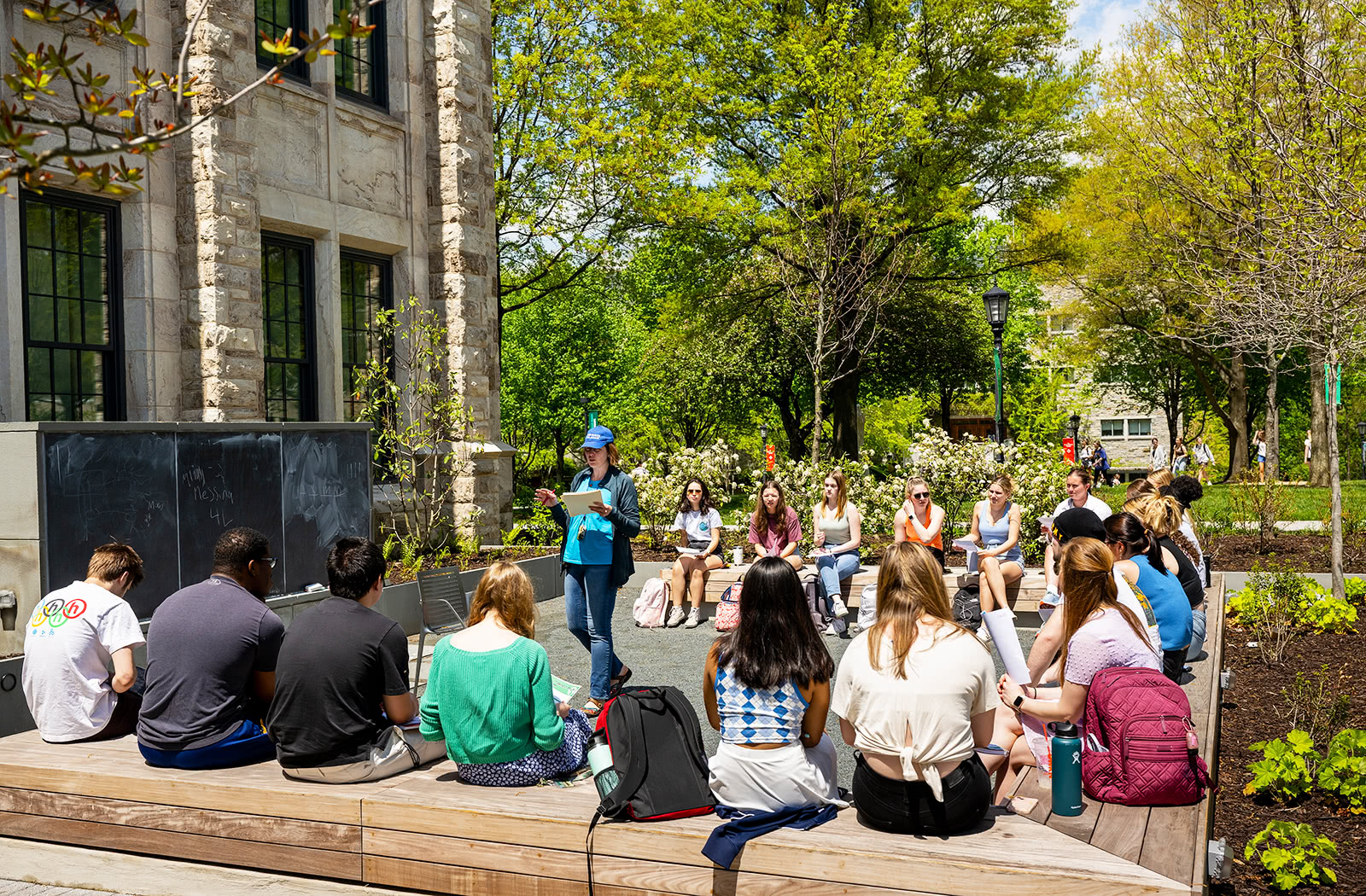 A group of students sitting on benches outdoors on a bright and sunny day watching a lecture