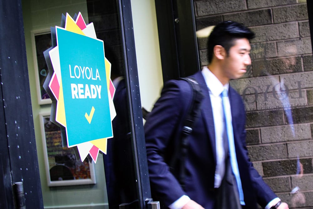 A student wearing a business suit walks out the door of a building with a sign that says 'Loyola Ready'