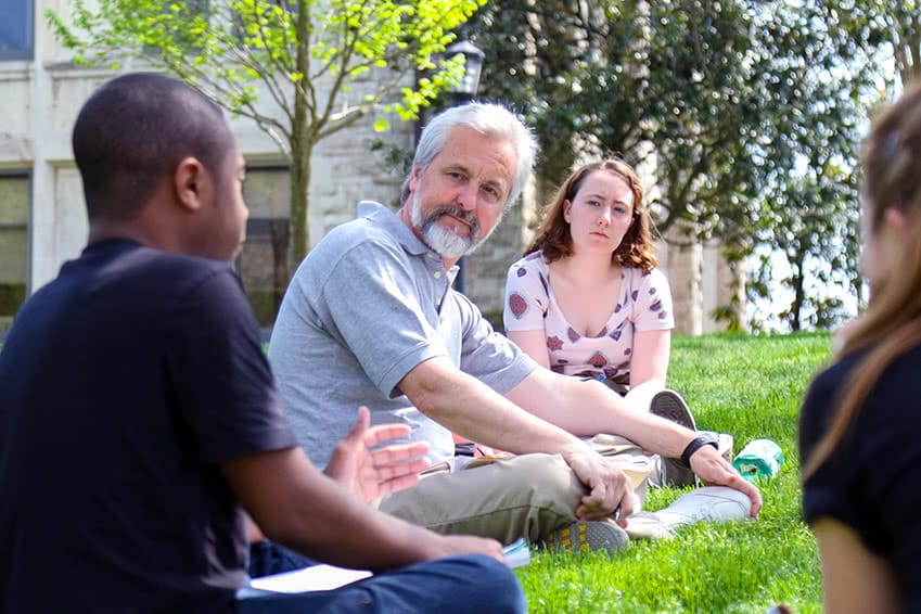 Students sit outside on grass with a professor having a discussion