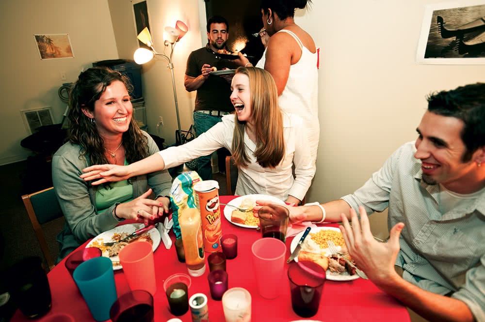 Students smiling and laughing around a table with a red tablecloth, food, plates, and drinks