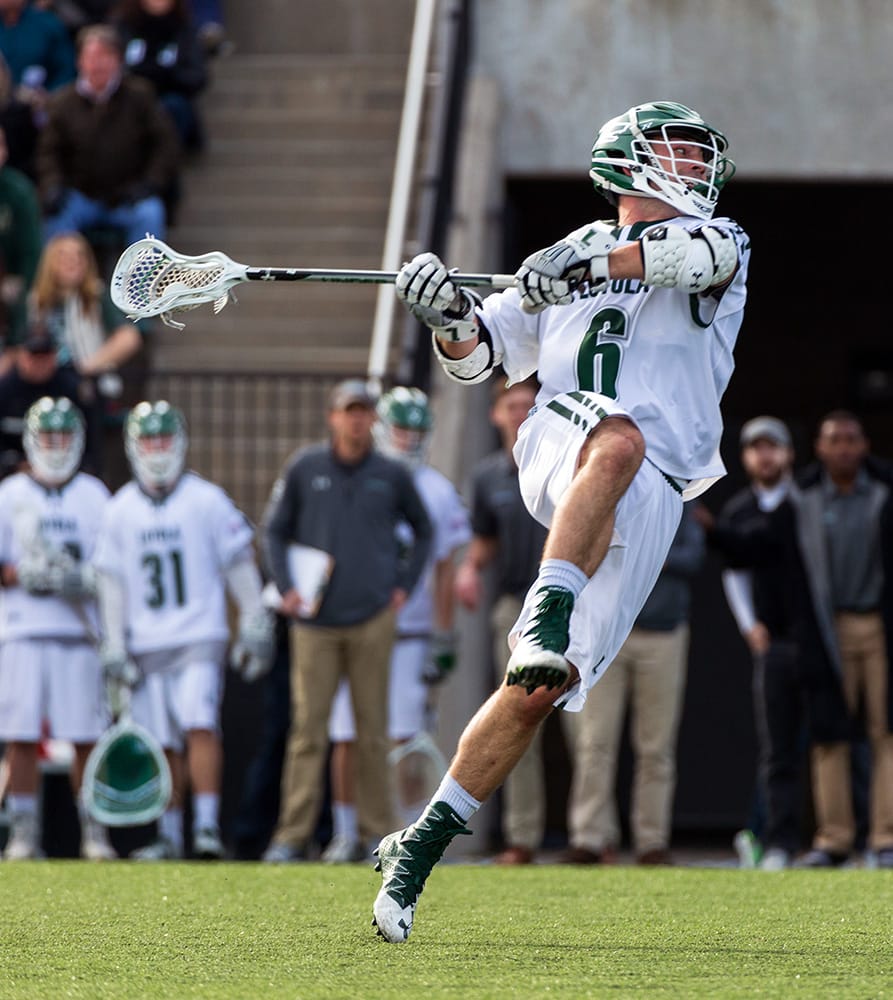 A men's lacrosse player swinging his stick about to throw the ball. Teammates can be seen in the background