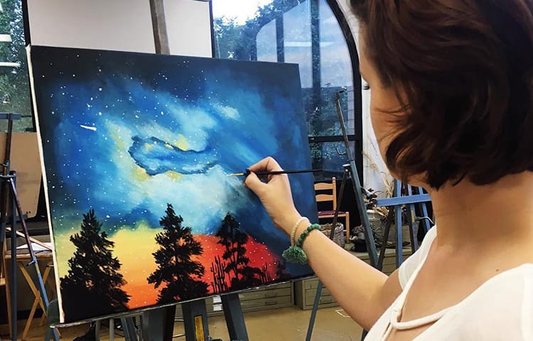 A student painting an outdoor scene with treetops and a glowing night sky