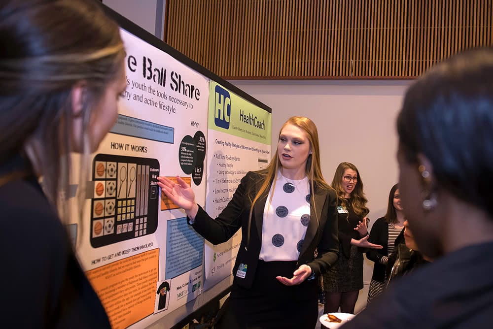 A student presents a poster while others watch and listen