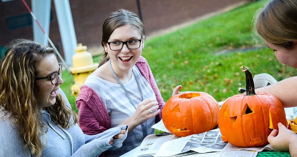 Three female students carving small orange pumpkins outside