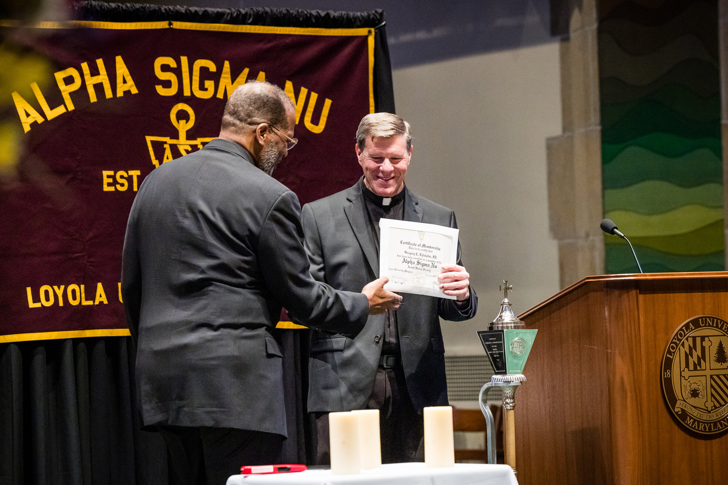 Two faculty members holding up an Alpha Sigma Nu Certificate