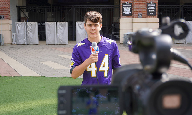 A young man wearing a Ravens jersey holding a microphone in front of a video camera