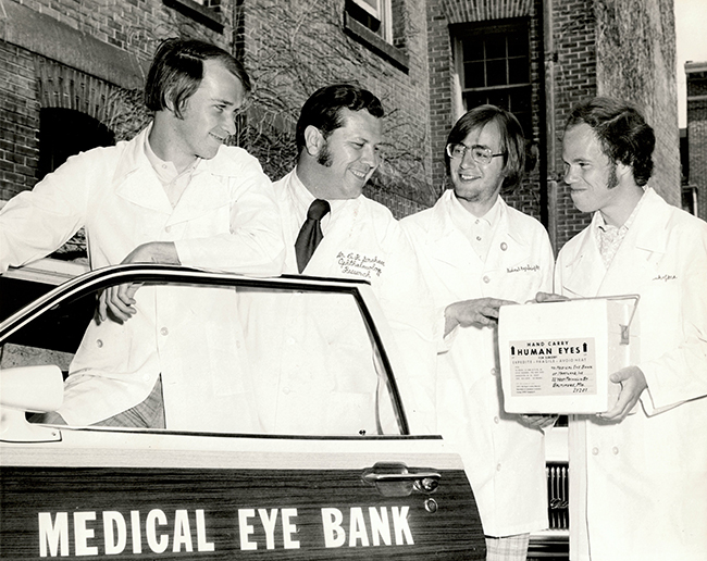 Black and white photo of Graham and three students preparing to transport a container of human eyes to the Medical Eye Bank for surgery. They are all wearing white lab coats and standing next to a car marked "Medical Eye Bank."