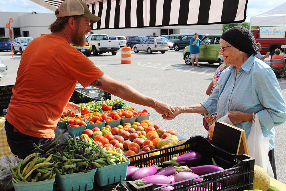 A woman purchases produce from a vendor at the Govans Farmers Market. The vendor has baskets of fresh produce on a table, including eggplants and tomatoes.